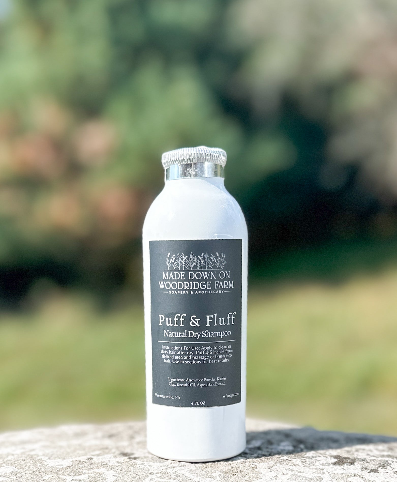 A white shaker bottle labeled 'Puff & Fluff Natural Dry Shampoo' placed on a rock with blurred greenery in the background - Naturally Linda