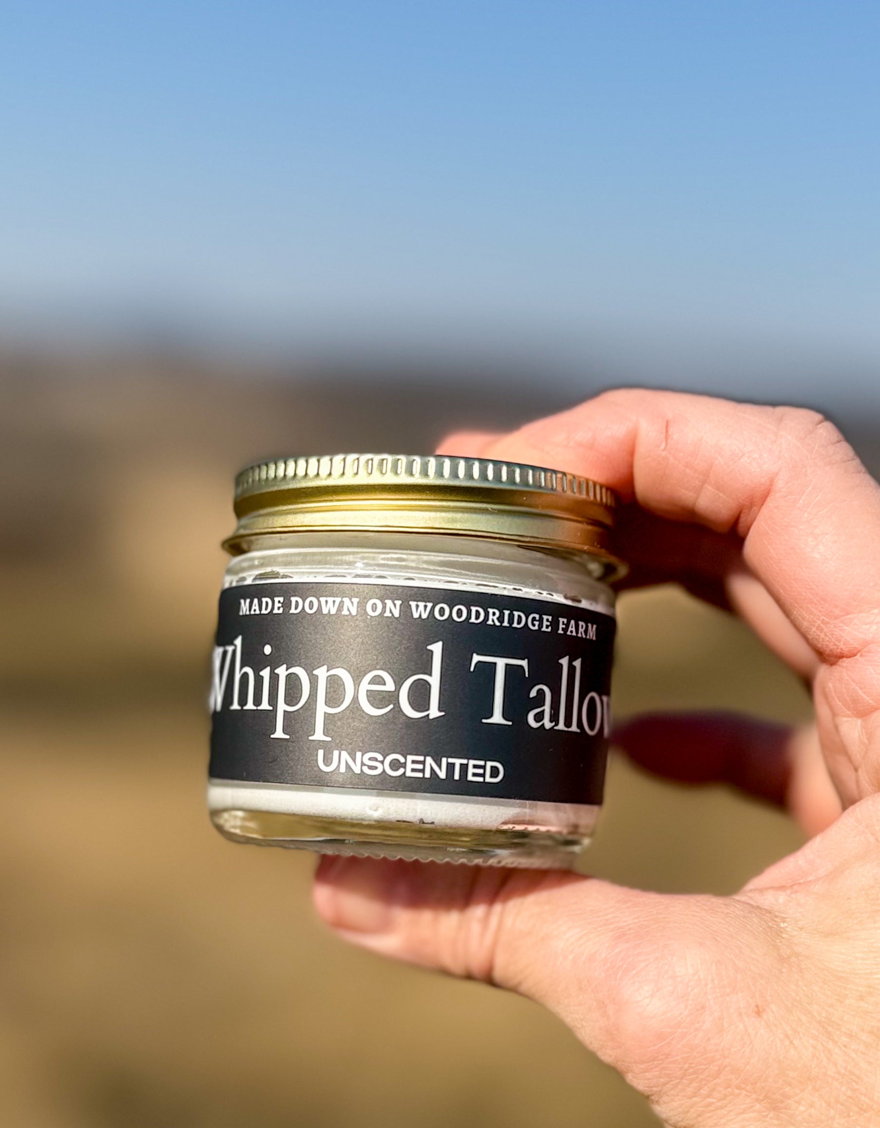 A jar of Whipped Grass Fed Tallow on a hand, with a vanilla bean flavor, against a grassy landscape background. - Naturally Linda