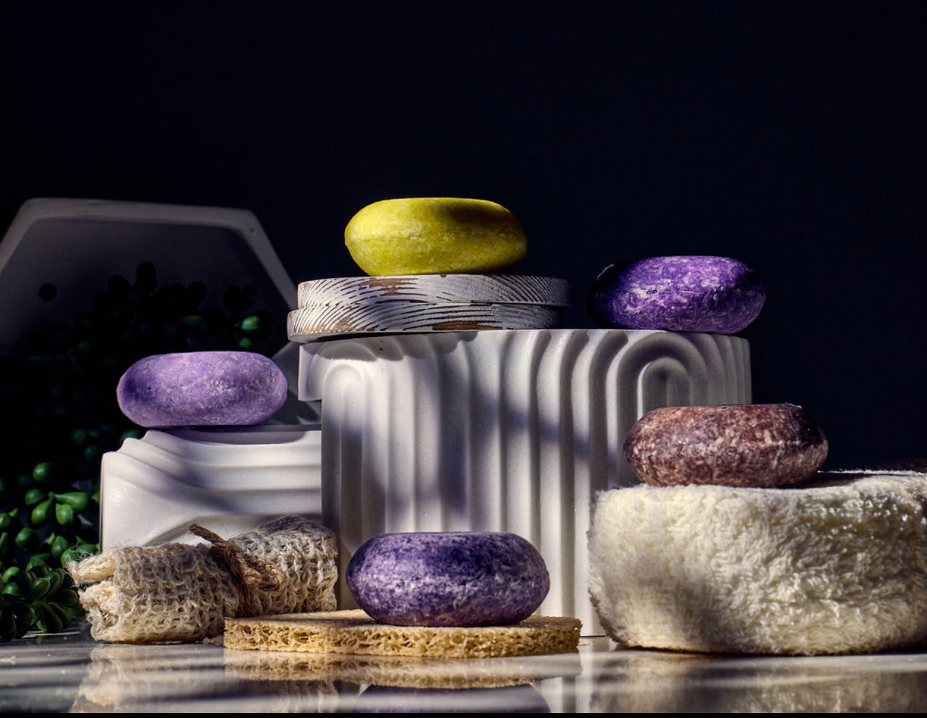 A variety of natural shampoo bars in different colors displayed on a marble surface with a white container in the background.