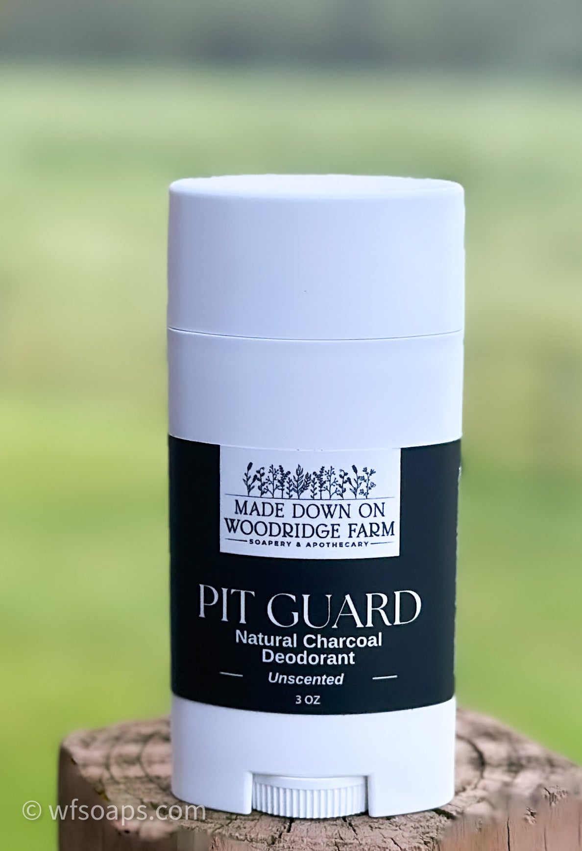 A white stick deodorant labeled 'PIT GUARD Natural Charcoal Deodorant' Unscented with a black lid, placed on a wooden surface with a green field in the background - Naturally Linda