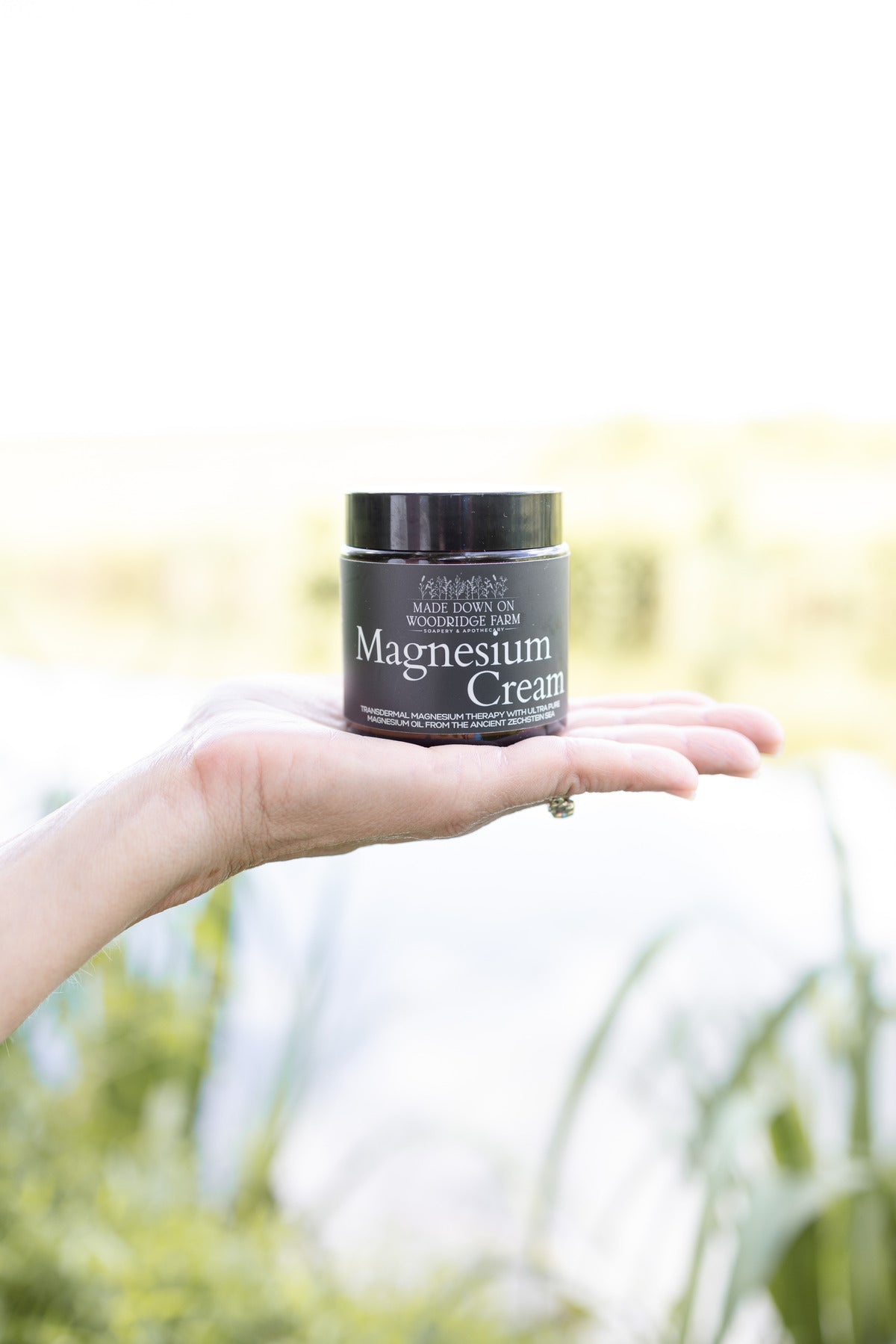 Hand holding a jar of magnesium cream with a blurred natural background - Naturally Linda