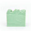 A handcrafted bar of soap with a greenish hue, placed on a wooden surface with a stone background.