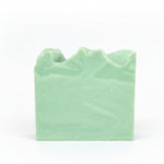 A handcrafted bar of soap with a greenish hue, placed on a wooden surface with a stone background.