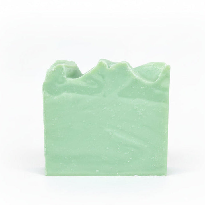 A handcrafted bar of soap with a greenish hue, placed on a wooden surface with a stone background.