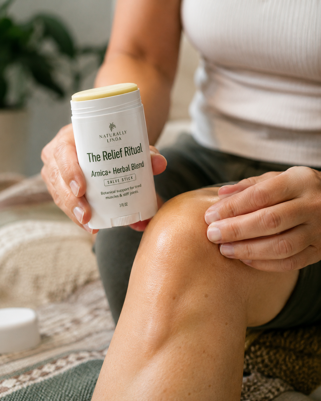 A person holding a white stick container labeled 'The Relief Ritual Arnica+ Herbal Blend' while touching their knee in a home setting.